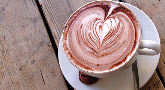 A steaming cup of hot chocolate with a heart-shaped swirl on top. Cup is placed on a wooden table. 